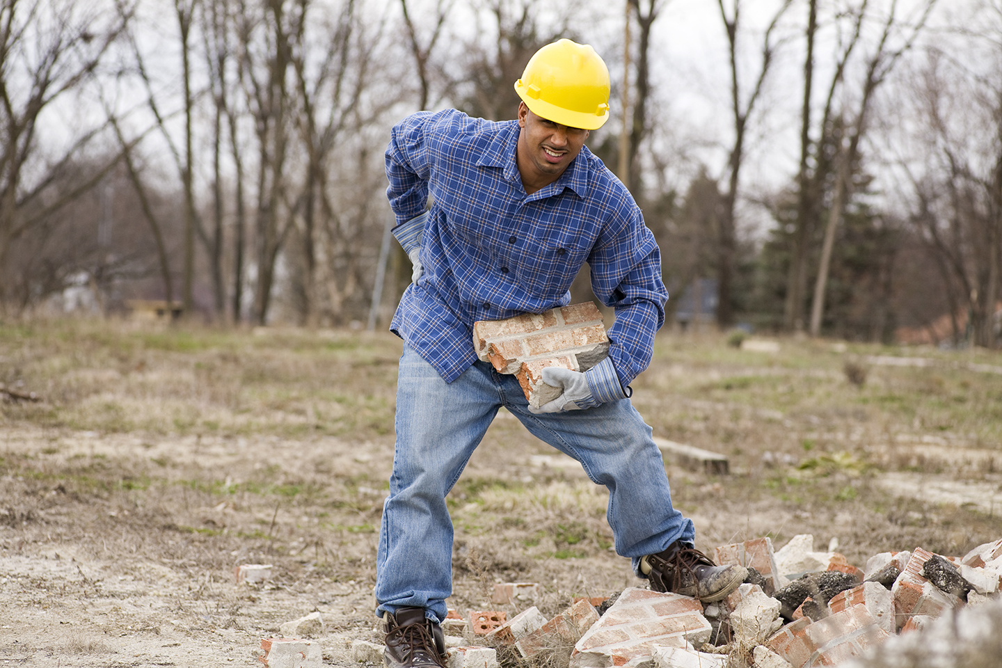 A worker in a hard hat and blue shirt carries bricks at a construction site.