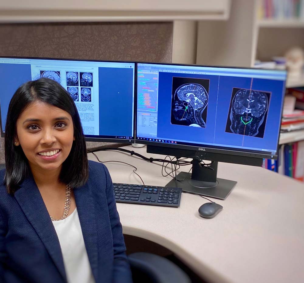 Lakshmi Kollara Sunil sitting at a desk with two monitors behind her showing MRI images of a brain.