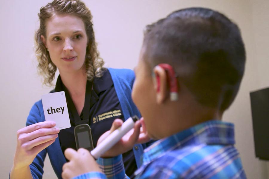 A woman showing a card that reads "they" to a young boy with a hearing aid on.