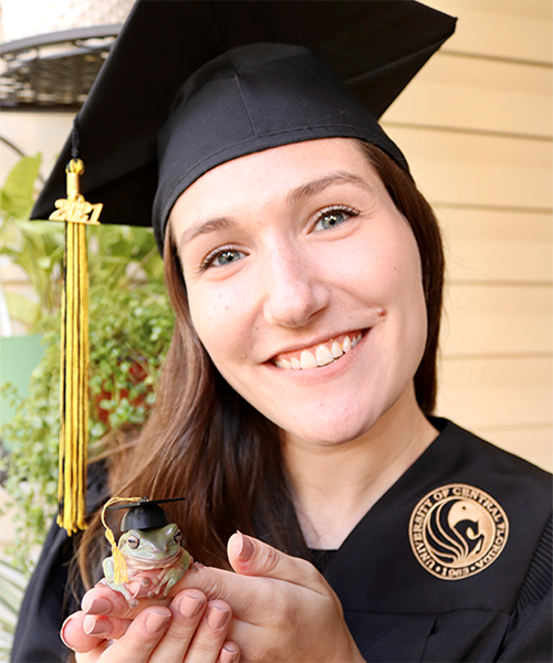 Kaitlyn Vidal holding a frog, smiling and wearing her UCF cap and gown.