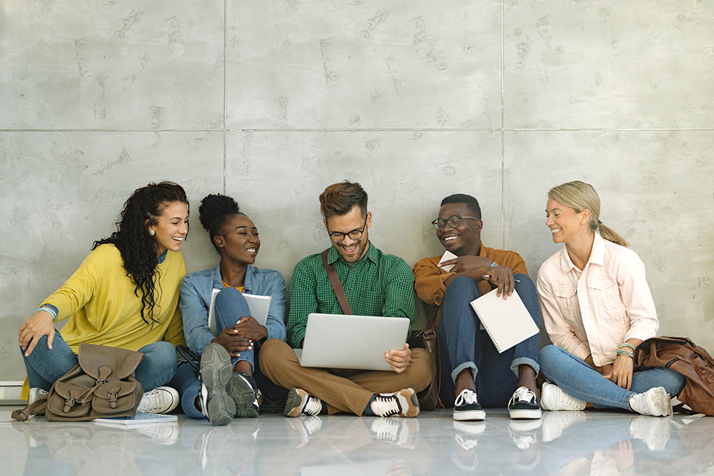 Group of five students sitting together, looking at a laptop and smiling.