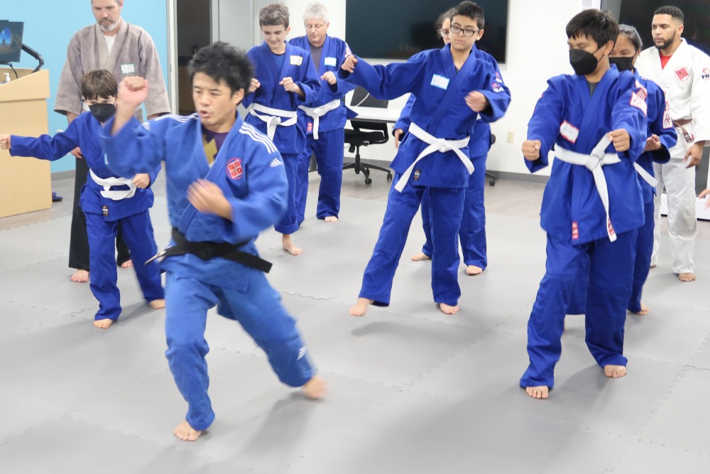 Adolescents in blue judogi (traditional uniform for Judo practice), practing judo techniques on a grey mat.