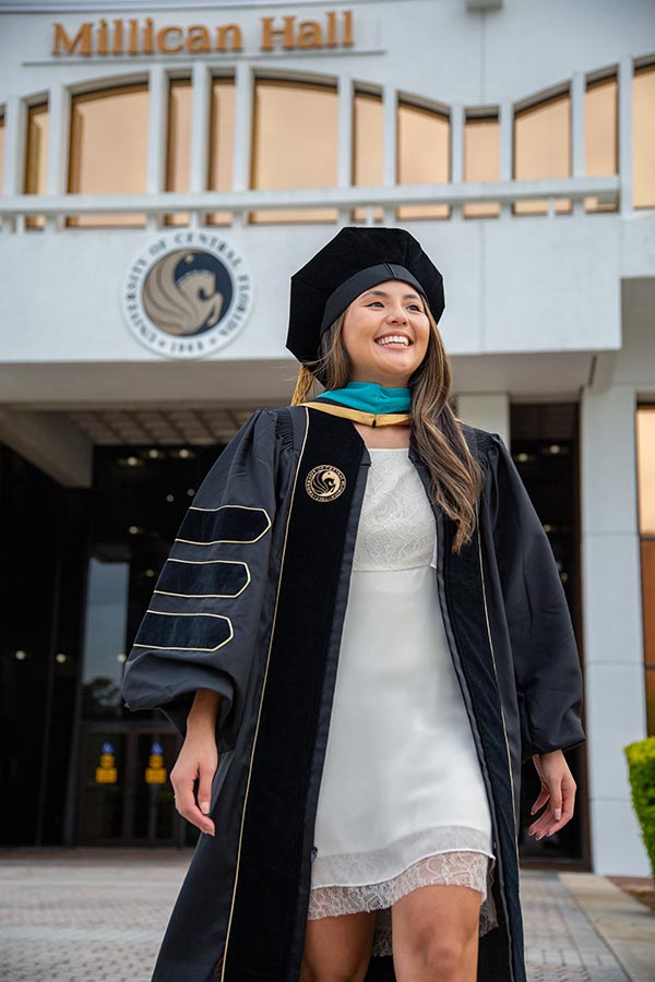 Mariah Bush walking in her graduation robe in front of Millican Hall on UCF's campus