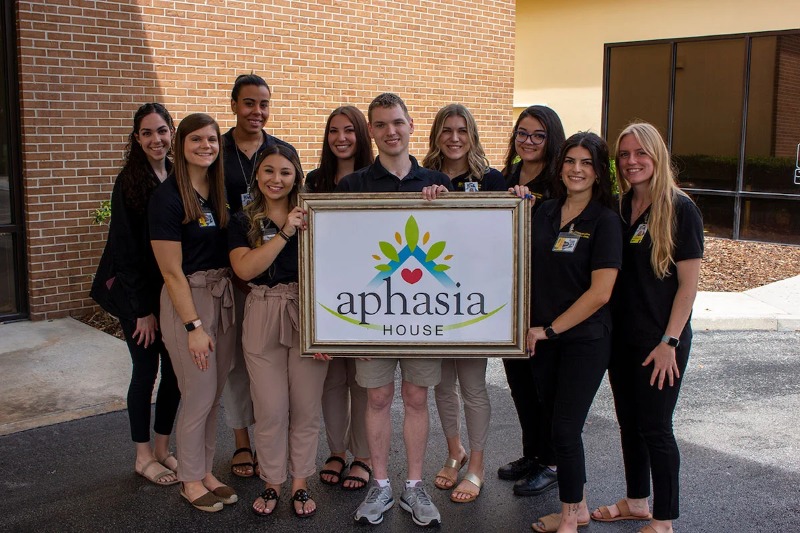 Kyle Burke (center) holding a picture frame with the Aphasia House logo while standing with graduate student clinicians.