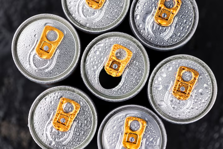 Aluminum cans with orange pop-tops are seen from above. There are six encircling one more in the middle which is popped open. There is water droplets on them, giving them a cold feel.