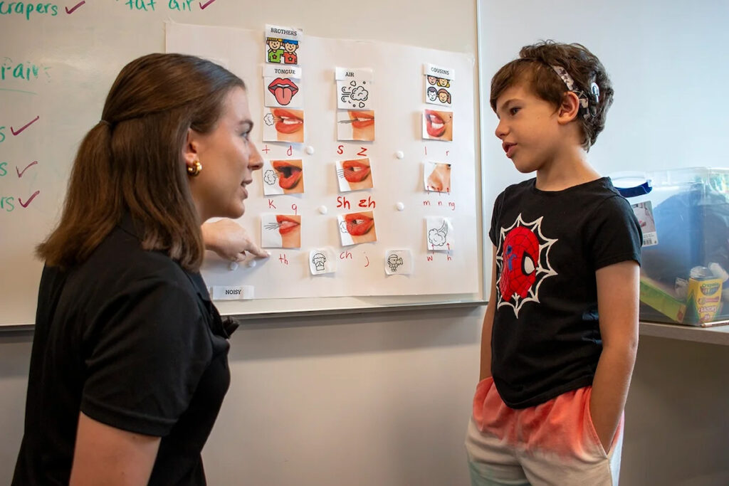 A woman speaking to a boy in front of a bunch of pictures on a white board.