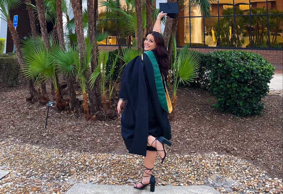 Fabiola Castro raising her graduation cap in the air while in front of palm trees.