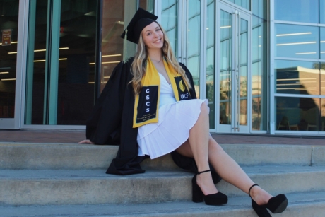 Hannalee Frahm wearing a white dress and black graduation cap and gown while sitting on steps.