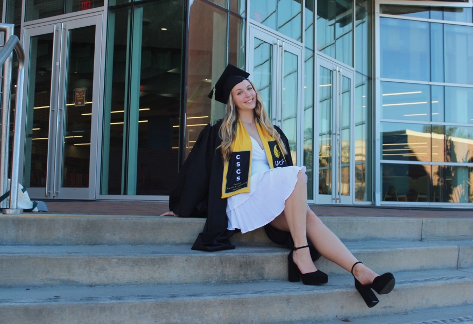 Hannalee Frahm wearing a white dress and black graduation cap and gown while sitting on steps.