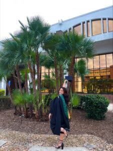 Fabiola Castro raising her graduation cap in the air while in front of palm trees.