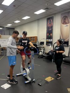 Fabiola Castro working with two students in a gym facility, with one student using a stationary bike. 