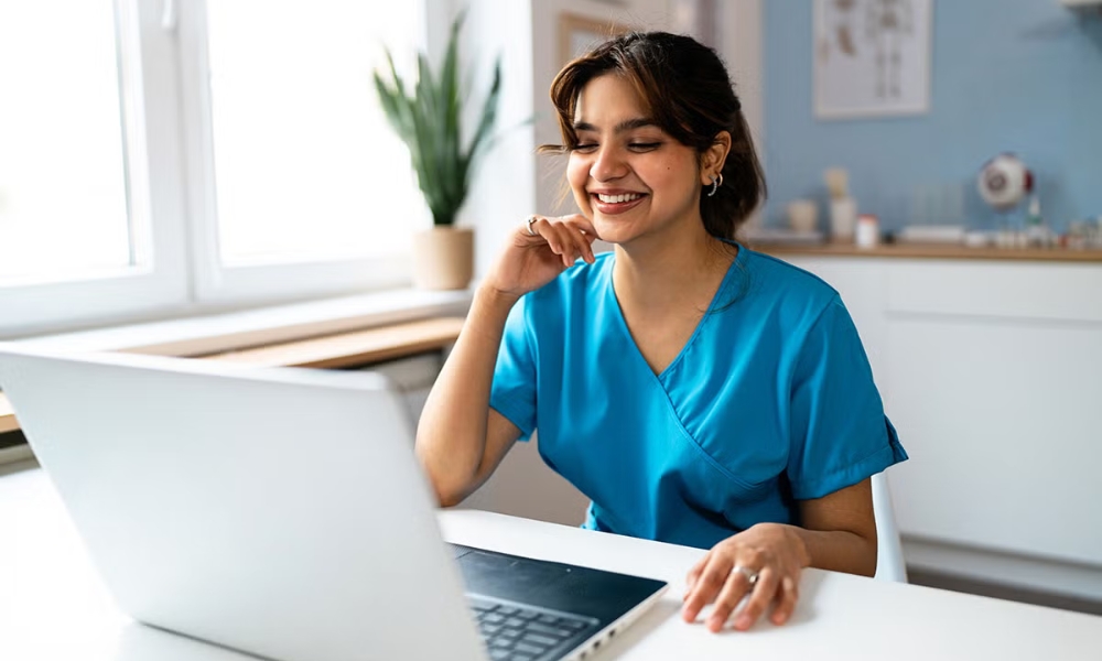 A healthcare worker sits in from of her laptop wearing blue scrubs.