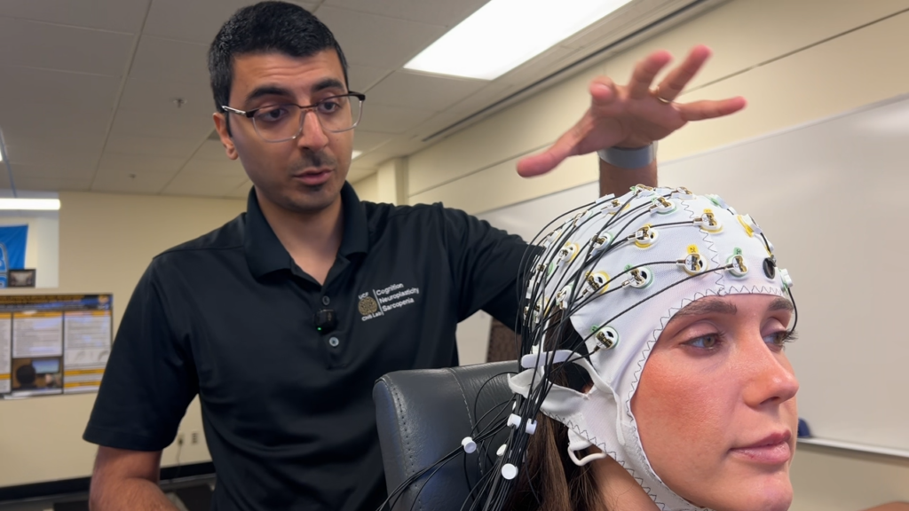 Moein Koohestani gestures above a seated assistant wearing an EEG cap fitted with multiple electrodes and wires, as he explains or prepares the equipment in a lab setting. The assistant looks forward while the connected sensors and cables extend from the cap, indicating brain activity monitoring.